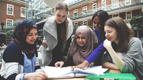 A diverse group of young women sitting around a table, books and laptops open, engaged in studying together.