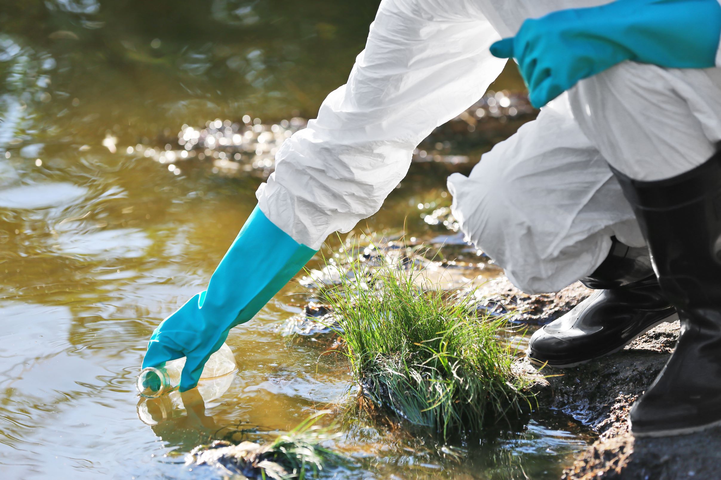 a person in white hazard suit with blue gloves reaches into the water to take a sample