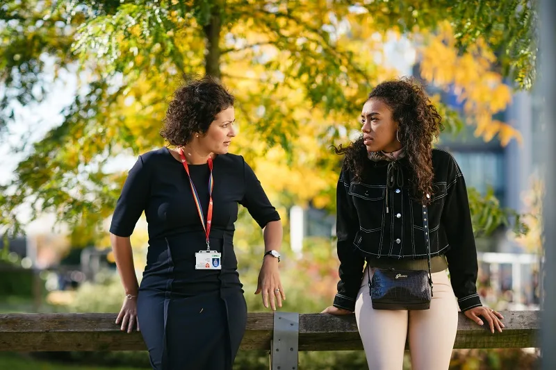 Two women, staff and students, engaged in conversation on a bench in front of lush green trees.