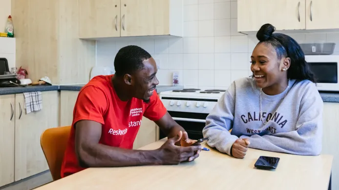Two young individuals sitting at a kitchen table, engaged in conversation with smiles on their faces.