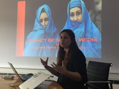 A reporter presenting to an audience with a screen displaying women in a blue scarf.