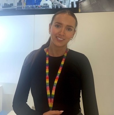 A young white woman with long hair tied back, in a black top and with a rainbow lanyard around her neck is smiling at the camera