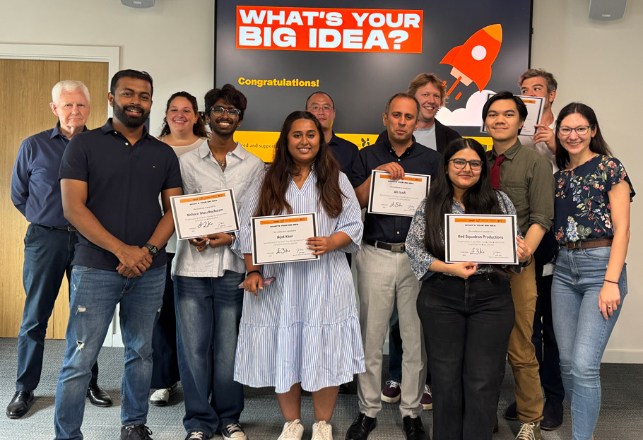 A large group of men and women stand, some of them holding certificates, in front a screen with the caption 