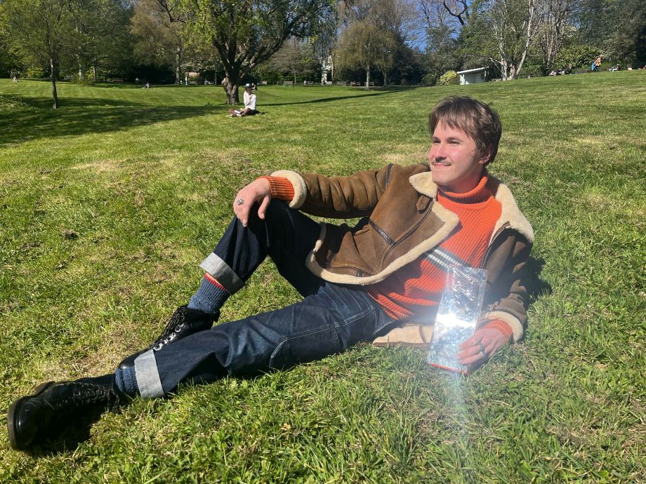 A young man in a sweater, sheepskin jacket and jeans reclines on a grassy slope holding a trophy made of clear plastic which glints in the sunlight
