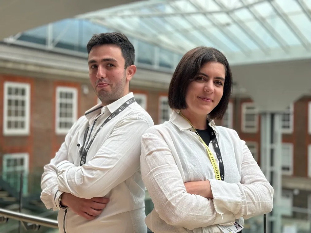 A young man and a young woman in identical white shirts and lanyards stand side by side with their arms crossed