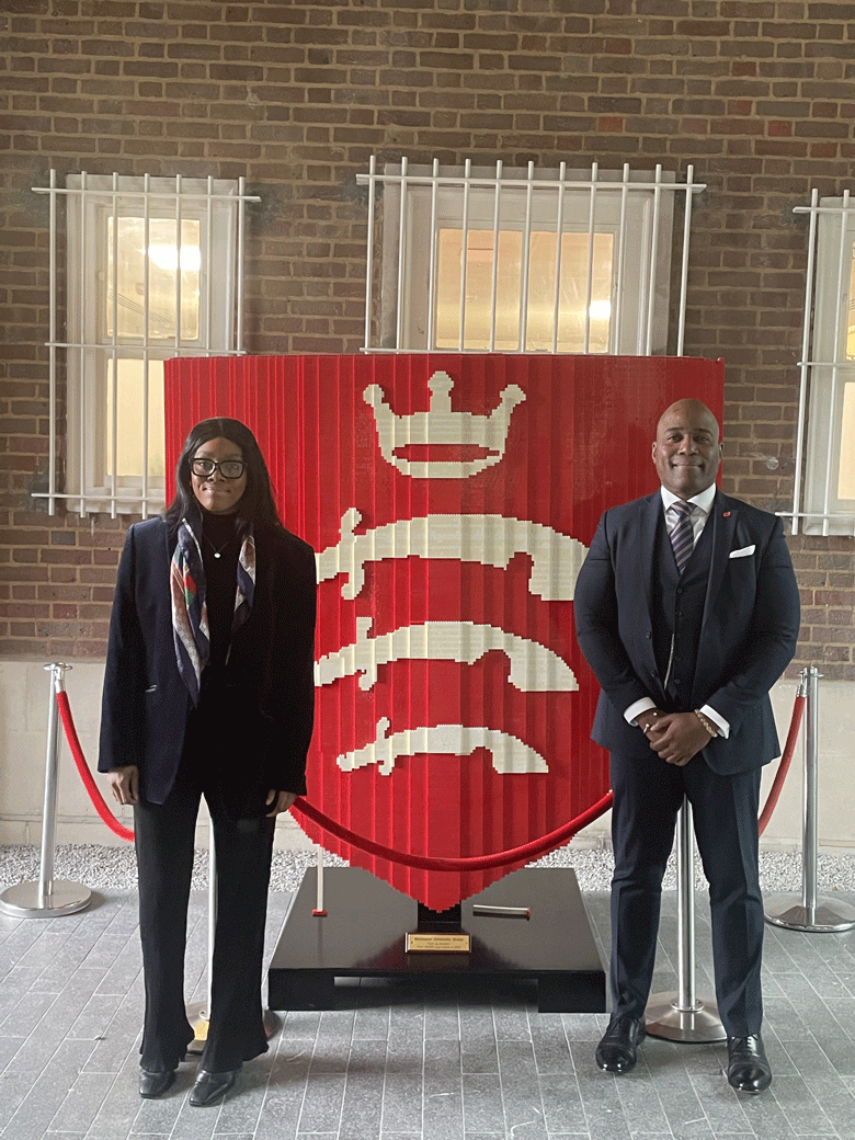 Two Nigerian visitors to MDX, in smart business wear, stand by the lego MDX shield in the entrance area of the College Building
