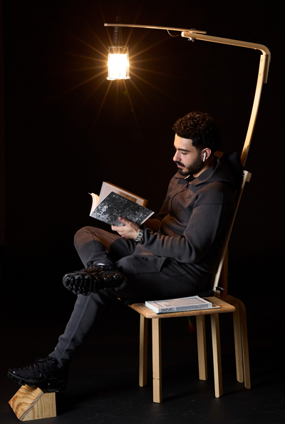 A male sitting on a chair reading with a light suspended above him