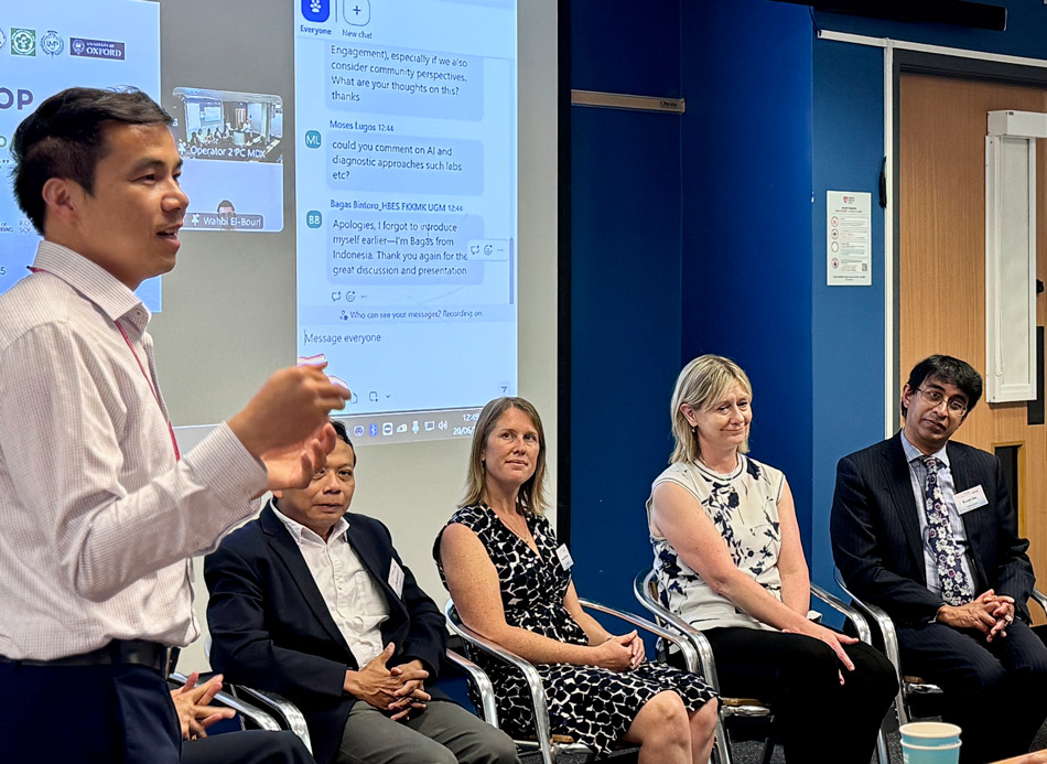 A panel of male and female speakers seated at a workshop. A male speaker stands on the left hand side of the picture, in the middle of addressing the audience