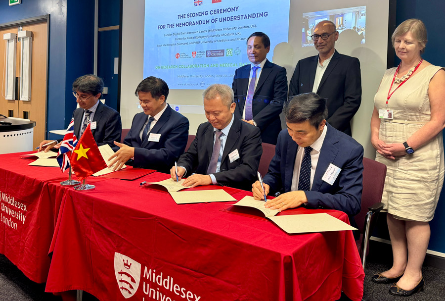 Four men in suits sit at a table with a red tablecloth on, signing documents. Two men and a woman stand behind them
