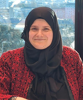 A woman with a black headscarf in front of a window smiles at the camera