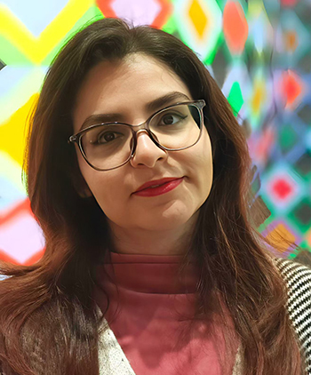 A woman with long hair and glasses looks at the camera in front of a colourful background