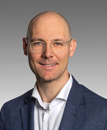 A man wearing a jacket but no tie smiles at the camera