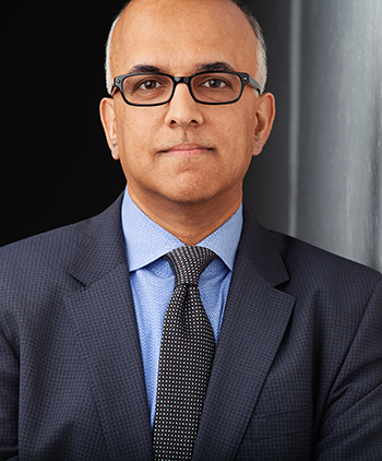A man in a jacket and tie with glasses looks straight at the camera