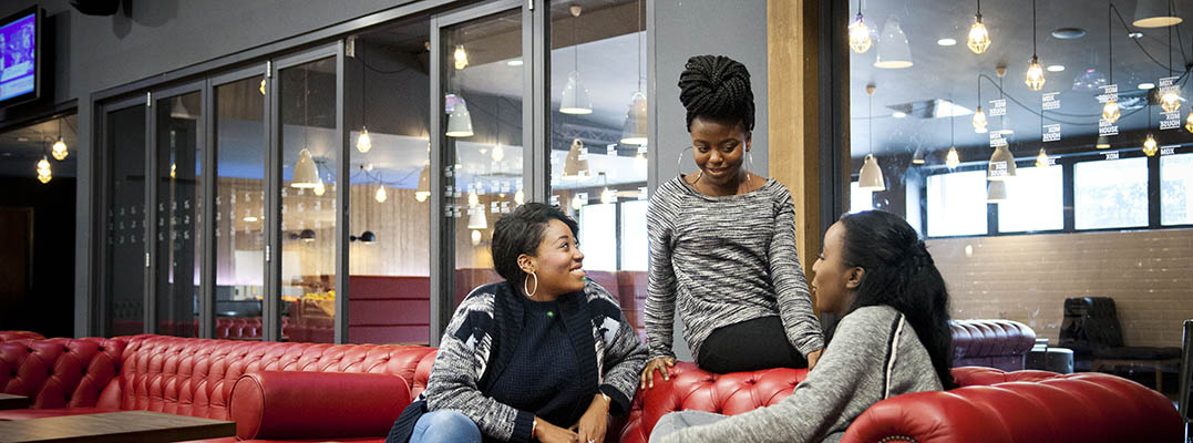 Three young women sitting on a red couch in a restaurant, captured in a Life on campus banner.