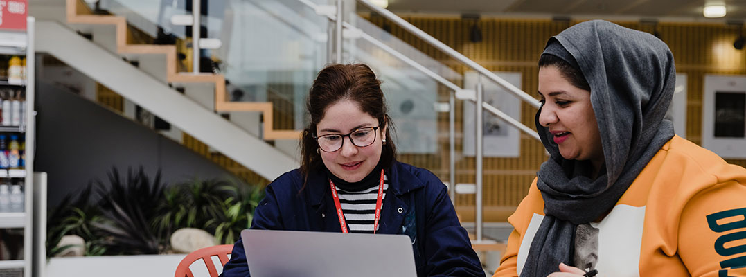 Two women sitting at a table with a laptop, discussing work projects and sharing ideas.
