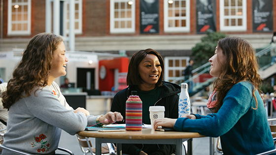 Three women enjoying a conversation at an outdoor cafe, sipping beverages and surrounded by greenery.