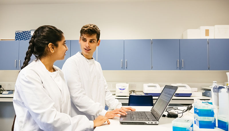 Two students wearing white coats in lab using laptop
