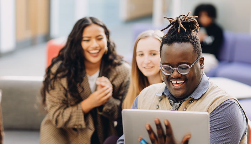 Student stood holding laptop which fellow students in the background.