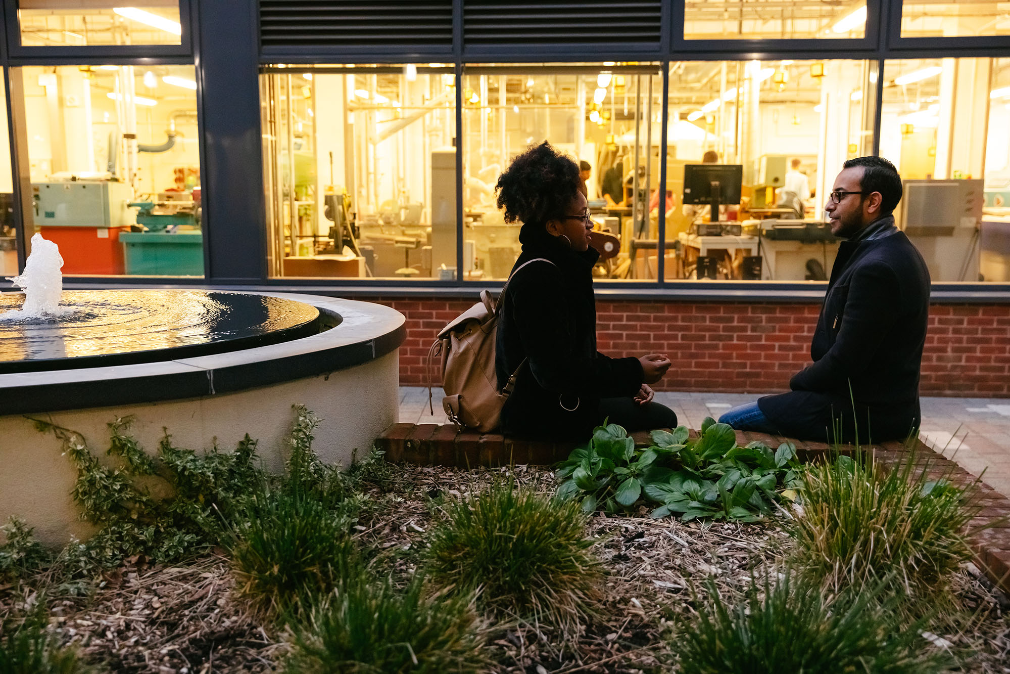 two students sit talking outside lit up building in early evening