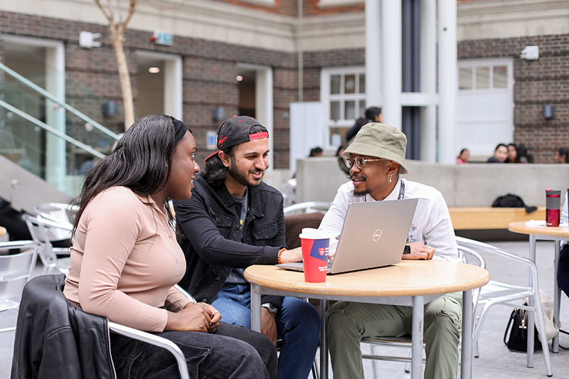 Three students collaborating on a project at a table with a laptop.
